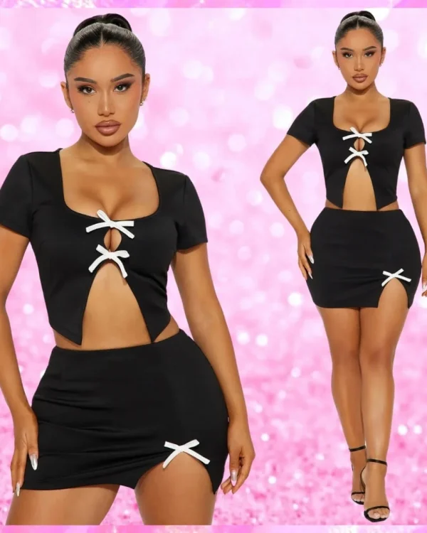 A woman poses in a black two-piece outfit with white bow accents against a pink, sparkly background. The outfit features a crop top and a mini skirt.